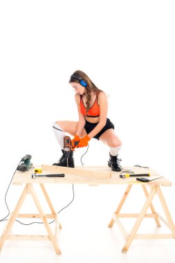 seductive girl in protective headphones working with electric drill at wooden table with tools, isolated on white clipart