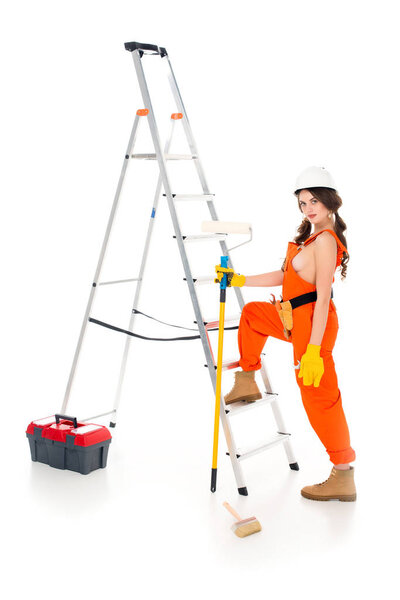 beautiful workwoman holding painting roller near ladder and toolbox, isolated on white