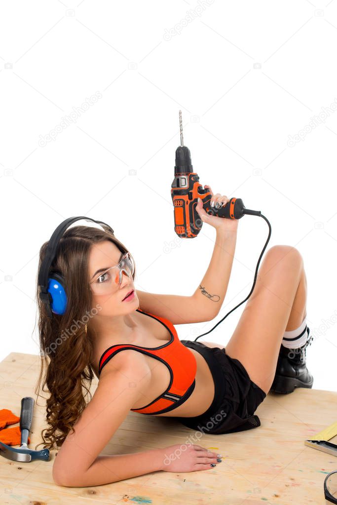 sexy girl in protective headphones posing with electric drill at wooden table with tools, isolated on white