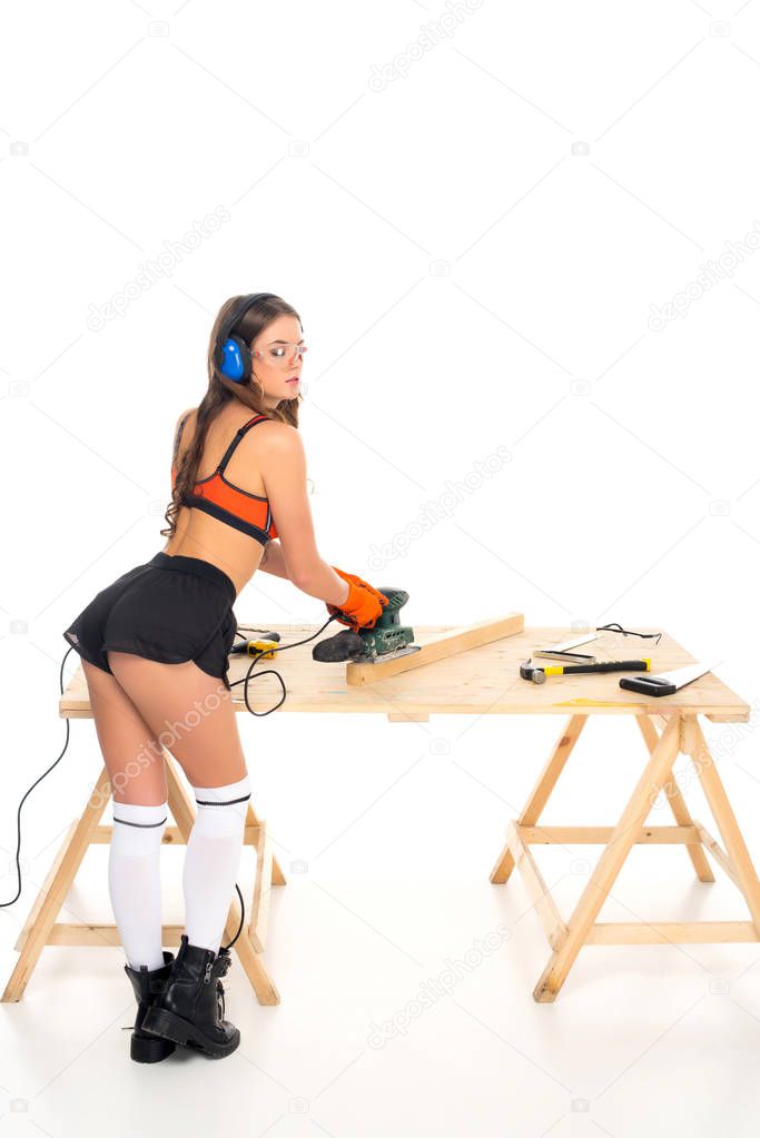 sexy girl in protective headphones working with grind tool on wooden table with tools, isolated on white