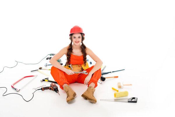 девушка в комбинезоне сидит на полу с различным оборудованием и инструментами, изолированные на белом
