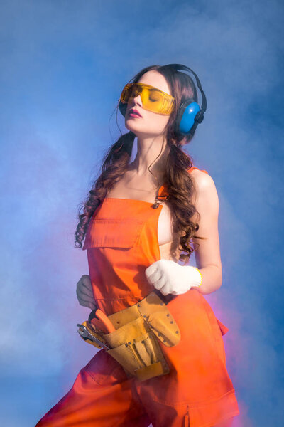 sexy girl in overalls with tool belt, goggles and protective headphones, isolated on blue with smoke