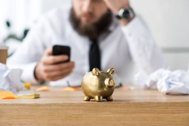 selective focus of piggy bank and businessman with smartphone clipart