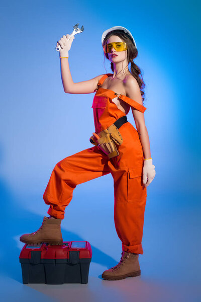 beautiful girl in overalls with tool belt and toolbox holding wrench, on blue