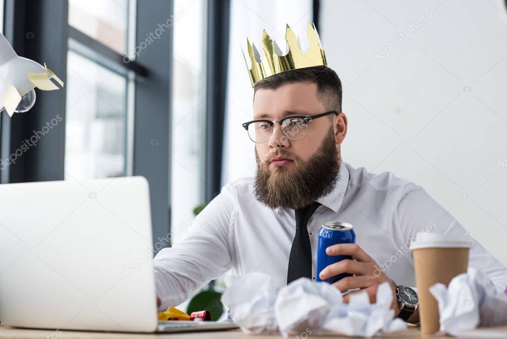 portrait of businessman with paper crown on head and soda drink in hand working on laptop at workplace in office