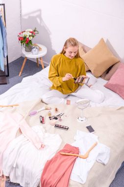 high angle view of young woman opening eye shadows box while sitting on bed clipart