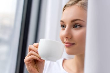 smiling young woman drinking coffee and looking through window clipart