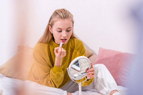 attractive young woman applying lipstick on bed at home