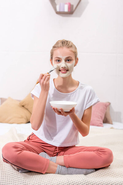 happy young woman applying clay mask on face at home and looking at camera