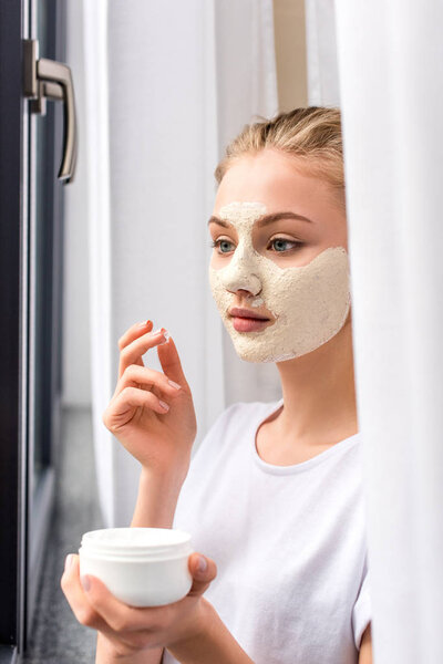 attractive young woman applying white clay mask on face at home