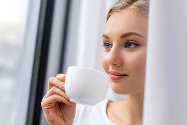 Leende Ung Kvinna Dricka Kaffe Och Titta Genom Fönstret — Stockfoto