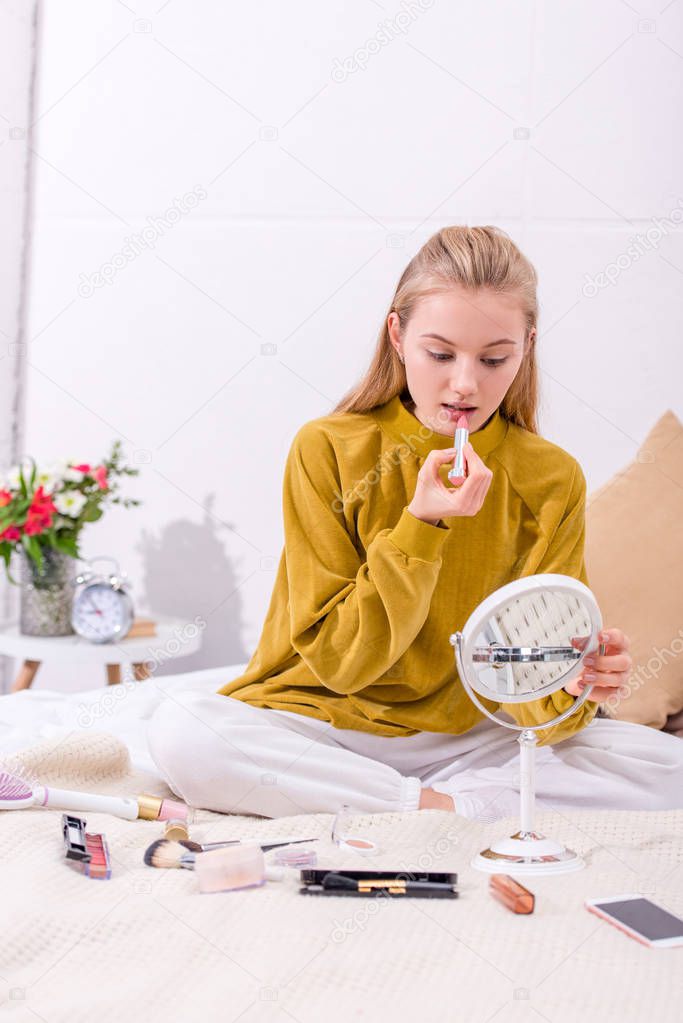 young woman applying lipstick on bed at home and looking at mirror