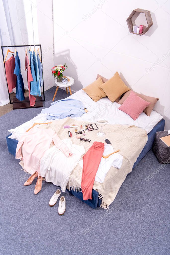 high angle view of interior of modern bedroom with hanger full of various female clothing and makeup supplies on bed