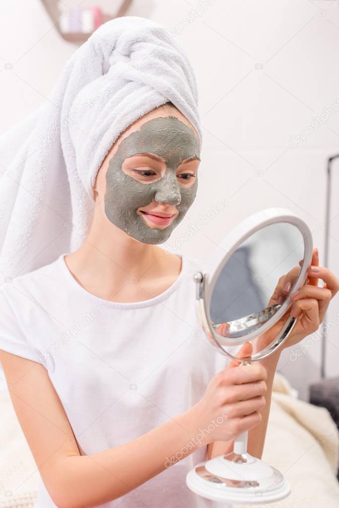 young woman with cosmetic clay mask looking at mirror at home
