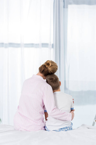 rear view of mother and son embracing while sitting on bed in morning