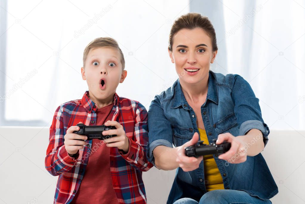 surprised mother and son playing video games with gamepads together and looking at camera