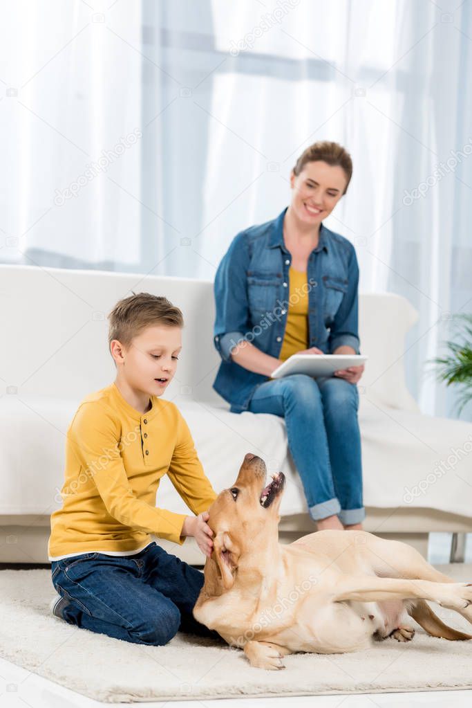 kid petting dog on floor while mother using tablet on couch