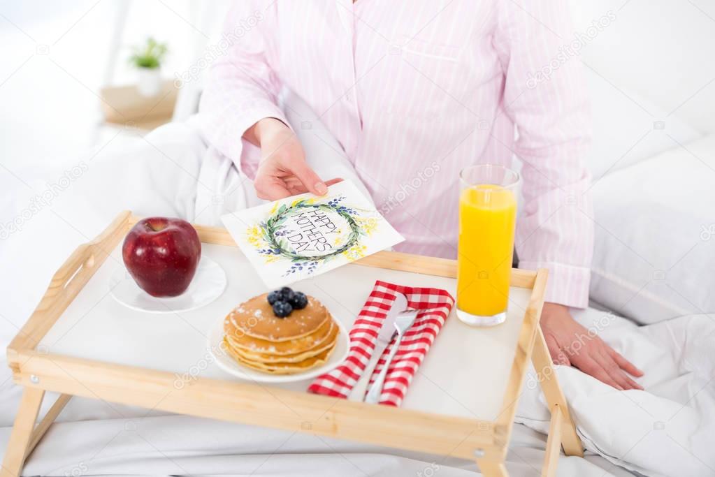 cropped shot of woman with breakfast in bed and mothers day greeting card