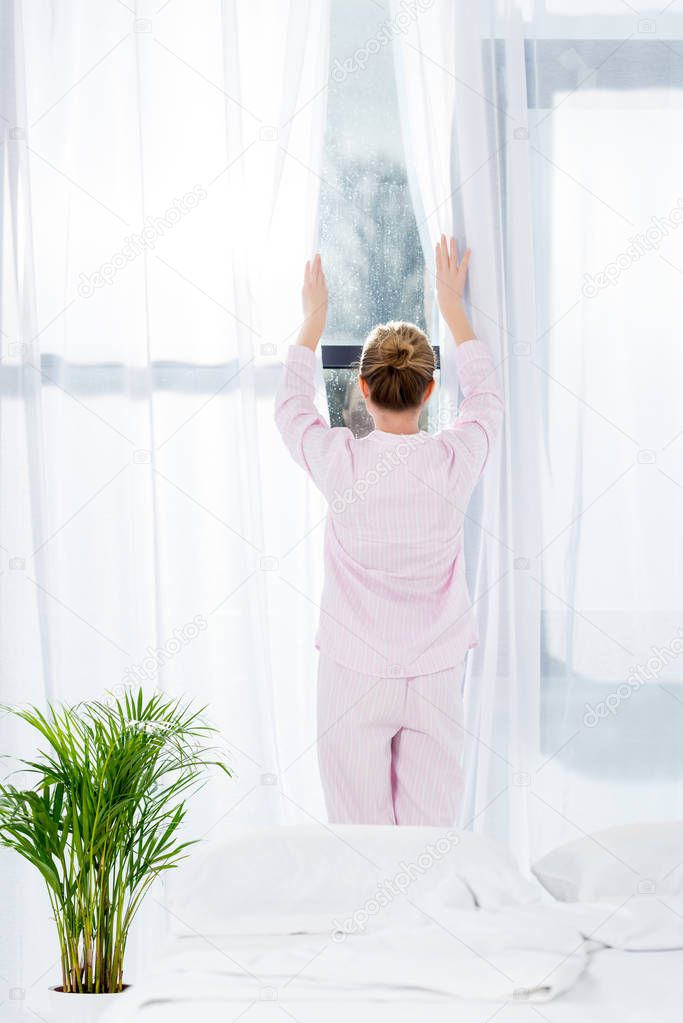 rear view of woman in pajamas opening curtains in morning