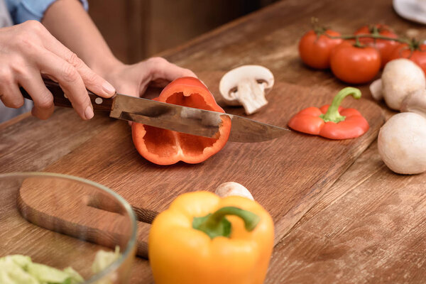cropped image of woman cutting red bell pepper on wooden board in kitchen