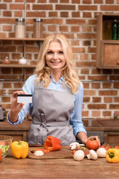 woman showing credit card and standing near table with vegetables in kitchen