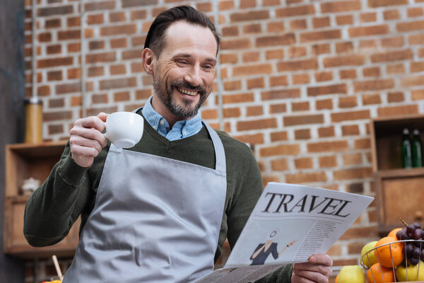 smiling man holding cup of coffee and reading travel newspaper