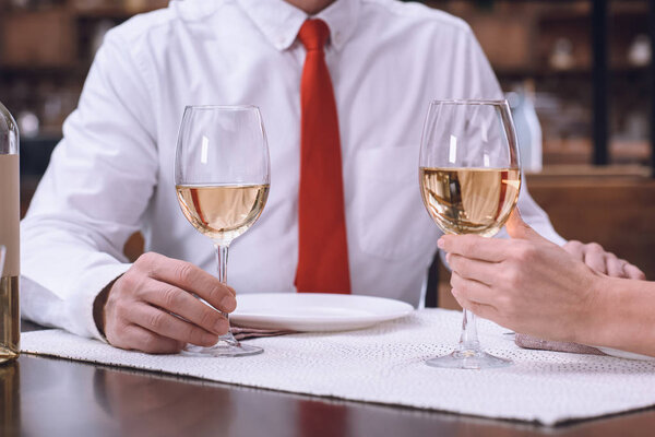 cropped image of couple holding hands and glasses of white wine at romantic dinner