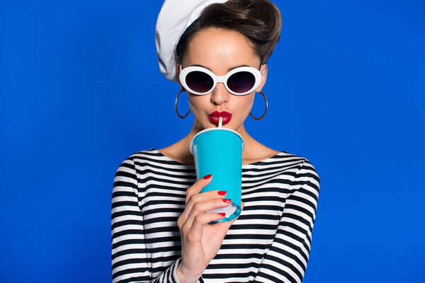 portrait of stylish young woman with beverage in paper cup isolated on blue