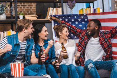 mutlu genç çok ırklı arkadaş kanepede otururken Amerikan bayrağı ve bira şişesi holding 