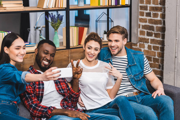cheerful young multiethnic friends taking selfie with smartphone while sitting on sofa