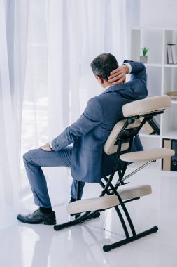 rear view of businessman with painful backache sitting in massage chair at office clipart