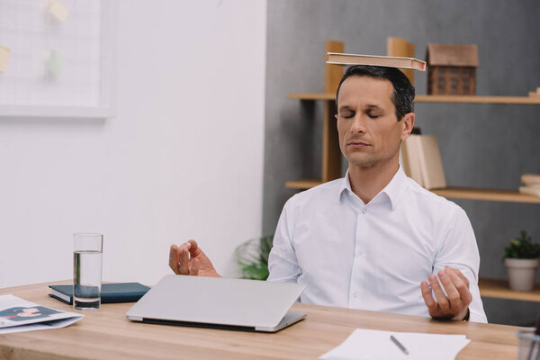calm businessman meditating with book on head at office