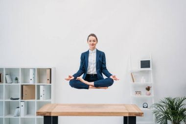 smiling young businesswoman with closed eyes meditating while levitating at workplace   clipart