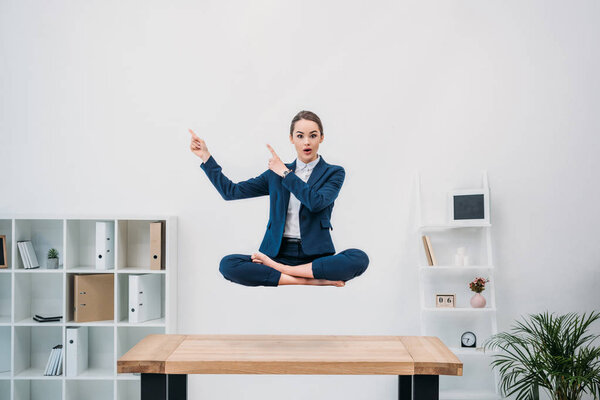 young busnesswoman pointing with fingers and looking at camera while levitating at workplace