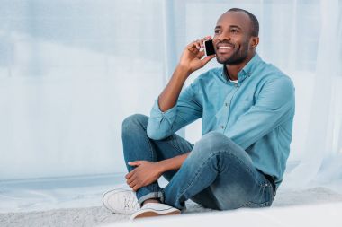 katta oturan ve evde smartphone ile konuşurken gülümseyen Afrikalı-Amerikalı adam