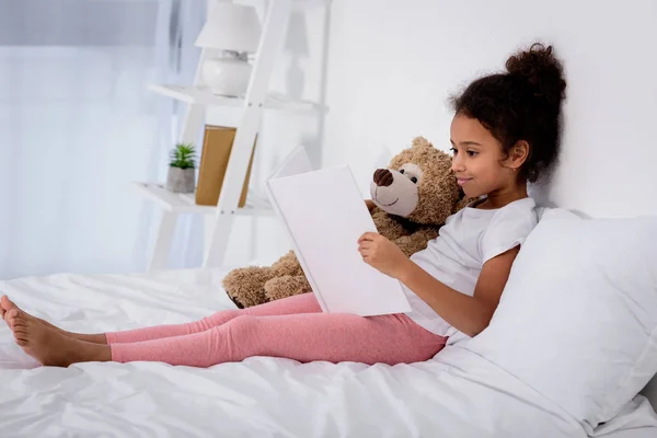 Adorable African American Kid Reading Book Hugging Teddy Bear Bed — Stock Photo, Image