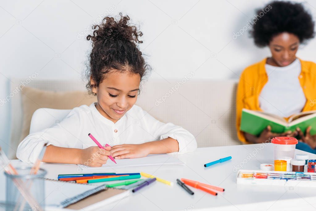 african american daughter drawing and mother reading book at home