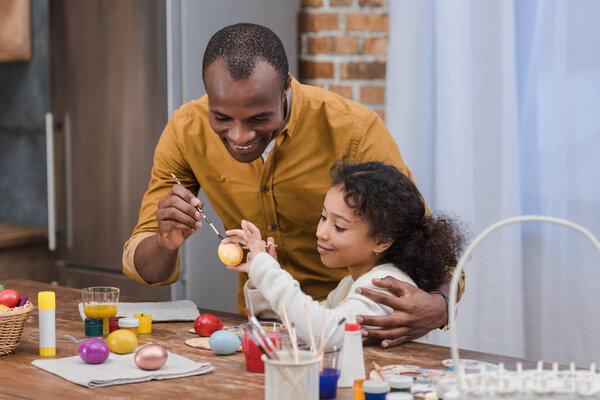 happy african american father and daughter painting easter eggs