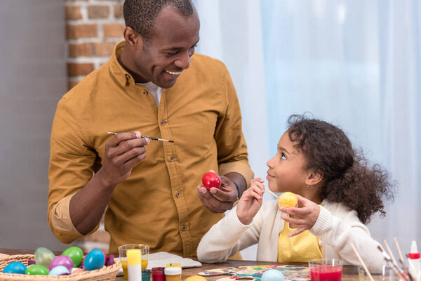 african american father and daughter painting easter eggs and looking at each other