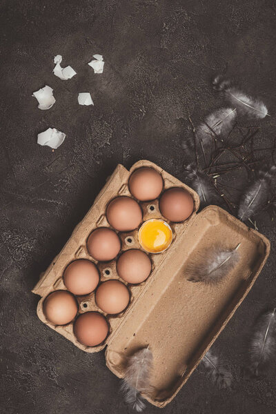 top view of chicken eggs and one broken egg with yolk in cardboard tray with feathers and branches on table