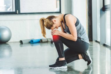 side view of young woman in sportswear having ache in knee while training in gym clipart