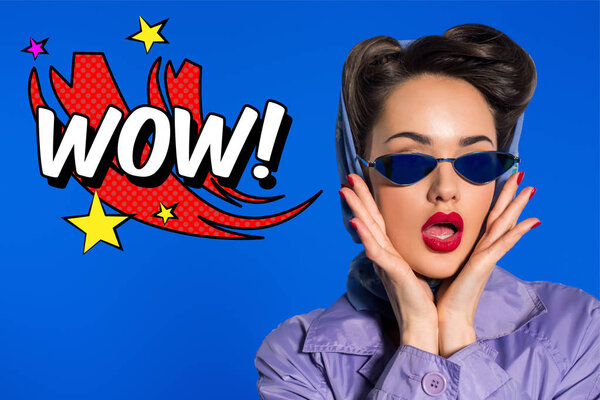portrait of stylish woman in retro clothing and sunglasses with comic style wow sign isolated on blue