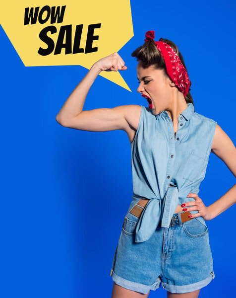 young woman in retro clothing showing muscles and shouting with sale speech bubble isolated on blue