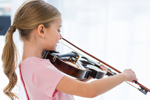 side view of cute little child in pink dress playing violin at home