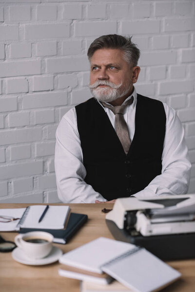 thoughtful senior writer sitting at workplace against white brick wall