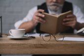 starší muž čtení knih na pracovní stůl s šálkem kávy a brýle na popředí