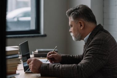 laptop ile çalışmaya ve Notlar not defterinde yapma düşünceli kıdemli yazar