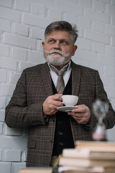 thoughtful senior man drinking coffee with stack of books and hourglass on foreground