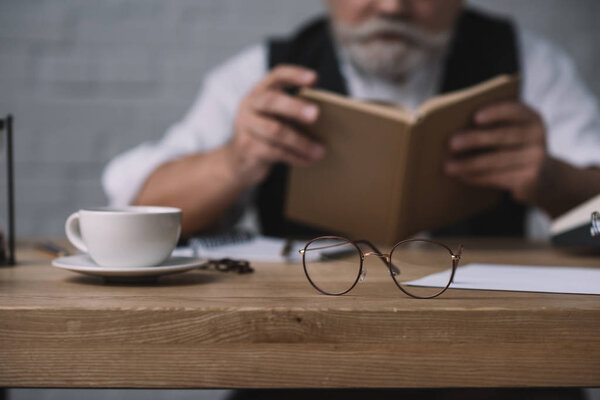 senior man reading book at work desk with cup of coffee and eyeglasses on foreground
