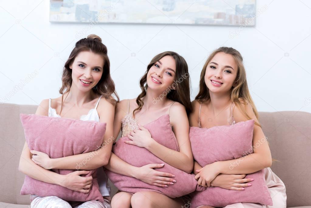 beautiful happy young women in pajamas holding pillows and smiling at camera